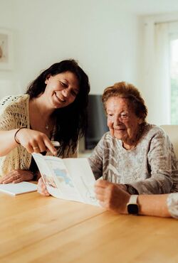 Seniorenbetreuerin sitzt an einem Tisch mit Seniorin und erklärt ein Formula